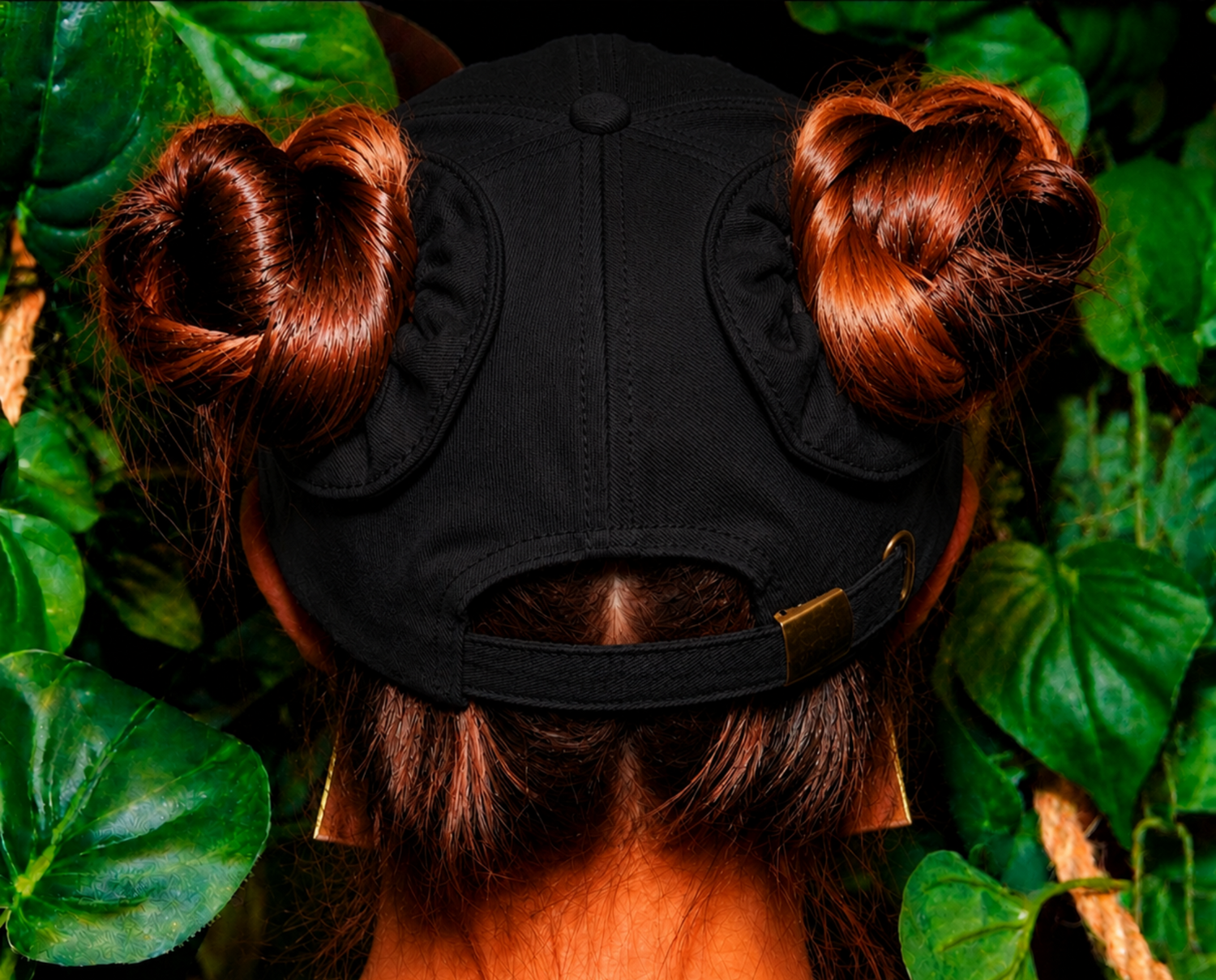 Person wearing a black cap or baseball hat with hair styled in space buns, surrounded by green leaves.